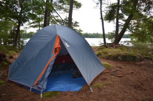 Large blue tent in pine forest beside a lake.
