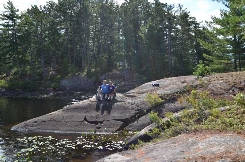 Two people and a dog on the rocks by a lake.