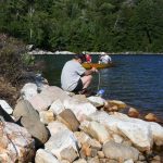 man on rocky shore pumping water, yellow canoe in bay